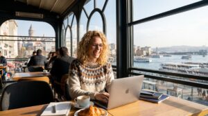 Working remotely in Istanbul | Monapart Hotel Mecidiyekoy Istanbul Woman with curly hair smiling while typing on a laptop in a sunlit cafe by a large window overlooking a river and cityscape.