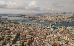 Aerial view of Istanbul with the Bosphorus, two bridges, boats, and a dense mix of low-rise houses and a distant high-rise skyline.